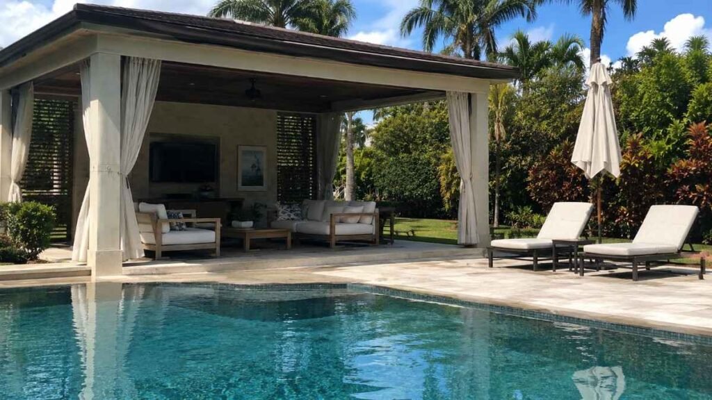 Luxury backyard with a swimming pool, featuring a covered pergola lounge area, outdoor seating, sun loungers, and tropical palm trees in bright daylight.